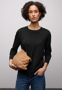 Pull noir en maille avec manches longues, col rond et poignets côtelés ; tenant un sac en daim beige. Jean bleu à coupe classique.