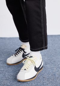 Vita Nike-sneakers med svart swoosh och gummisålar, bärs med vita strumpor och svarta byxor med vit söm, står på blå matta.