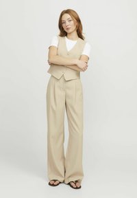 Beige vest met drie knopen, gedragen over een witte t-shirt, gecombineerd met losse beige broek en platte sandalen. Effen achtergrond.