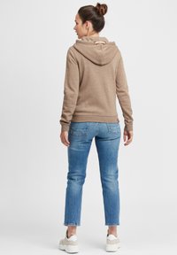 Brun hettegenser med en myk tekstur og snorhette, kombinert med lyseblå jeans og hvite sneakers. Bakfra viser den minimale designdetaljer.