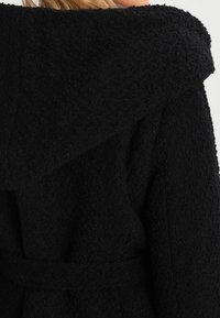 Mustat huppukevät takki, jossa on tuntuma, pörröinen kangas. Varustettu vyöllä vyötäröllä ja suurella kauluksella, mikä korostaa lämpöä ja mukavuutta.