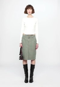 White long-sleeve crop top, olive green knee-length skirt with pockets, and knee-high black boots. Model holding a small burgundy handbag.