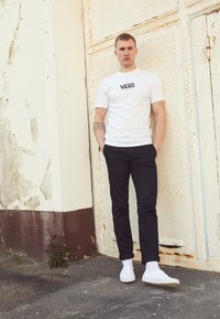 White short-sleeve Vans t-shirt with black logo, paired with black slim-fit pants and white sneakers. Textured cracked wall in background.