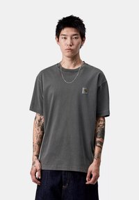 Giovane uomo con capelli neri corti, che indossa una maglietta grigia Carhartt, una collana in argento e jeans scuri, con numerosi tatuaggi sulle braccia e sul collo.
