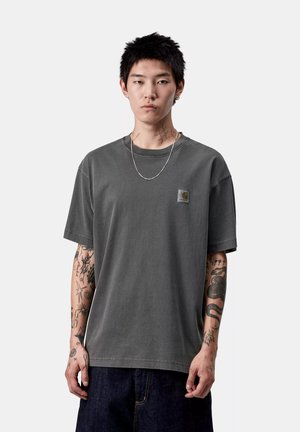 Giovane uomo con capelli neri corti, che indossa una maglietta grigia Carhartt, una collana in argento e jeans scuri, con numerosi tatuaggi sulle braccia e sul collo.