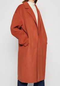 Donna che indossa un ampio cappotto di lana color ruggine sopra un maglione crema e jeans blu, in piedi con le mani in tasca contro uno sfondo semplice.