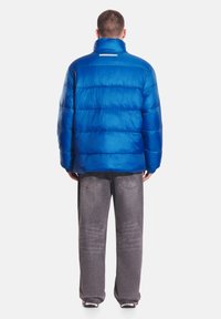 Niebieska puchowa kurtka z wysokim kołnierzem, pikowanym wzorem i poziomymi przeszyciami. Z tyłu znajduje się odblaskowy pasek. Połączona z szarymi jeansami.