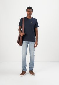 T-shirt bleu marine à manches courtes avec un logo, jeans en denim bleu clair, chaussures en cuir marron et sac en bandoulière en cuir marron.