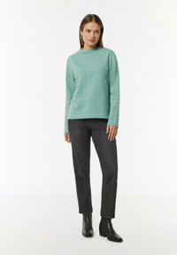 Mintgrüner, strukturierter Pullover mit hohem Kragen, getragen mit dunklen Jeans und schwarzen Ankle Boots, die einen lässigen Schnitt bieten.
