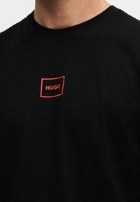 T-shirt noir avec un petit carré rouge contouré et le mot "HUGO" centré sur la poitrine sous l'encolure.
