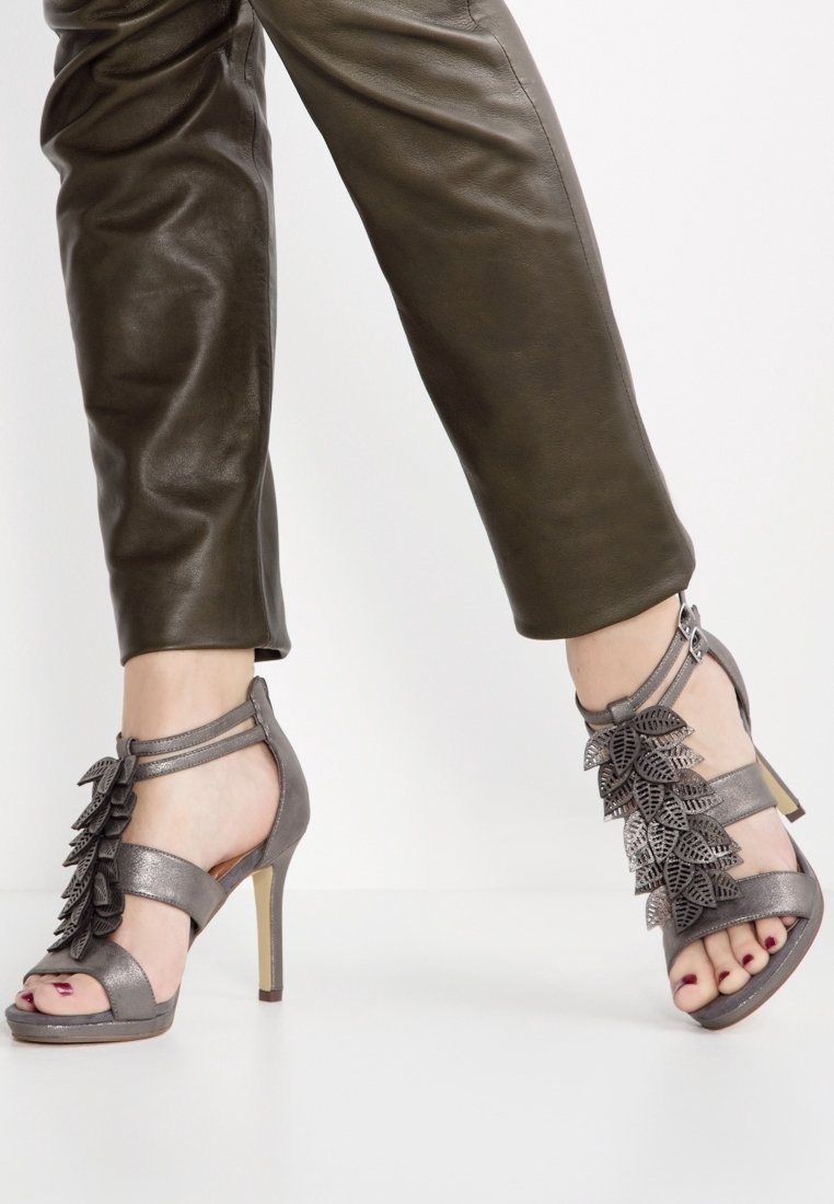 Femme portant un pantalon en cuir vert olive foncé et des sandales à talons hauts gris métallisé avec des ornements en forme de feuille et des brides à la cheville.