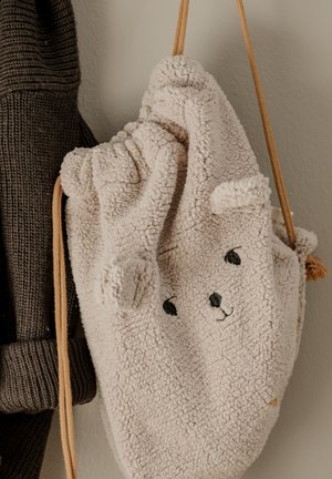 Bež plišana torba s ušima medvjedića i izvezenim licem, zgusnutim vrhom i smeđim vezicama za zatvaranje. Teksturirana, dlakava vanjština visi pored tamnog pletenog predmeta.