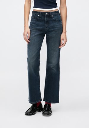Femme portant un jean large bleu foncé, des mocassins noirs brillants, des chaussettes rouges et un top court noir, debout devant un fond blanc uni.