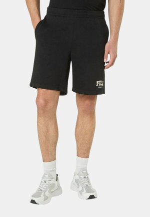 Uomo con pantaloncini neri FILA, calzini bianchi alla caviglia e sneakers bianche e grigie, in piedi con le mani rilassate lungo i fianchi.