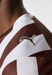 Stribet sporttrøje i brun og hvid med en tekstureret stof. Har et fremtrædende hvidt Puma-logo på skulderen.
