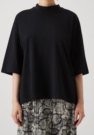 Osoba ubrana w gładką, czarną, oversize'ową koszulkę i plisaowane spodnie z wysokim stanem w czarno-biały wzór skóry węża.