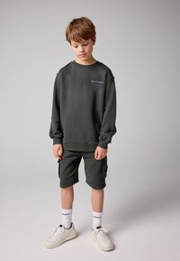 Mörkgrå sweatshirt och shorts i mjukt tyg, med en liten blå logga på bröstet. Matchas med vita sneakers och strumpor.