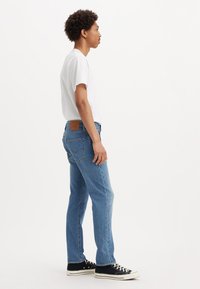 Jean en denim bleu clair à coupe droite, avec une patch en cuir marron, un design à cinq poches et une légère décoloration. Associé à des baskets noires.