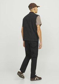 Black sleeveless jacket over a brown T-shirt, paired with dark jeans and brown sneakers with cream accents. The person faces away from the camera.