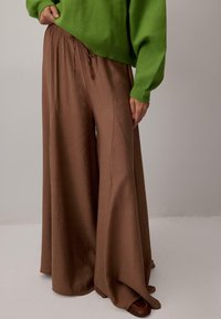 Rjave široke hlače z elastiko in vezalko v pasu, iz mehkega blaga, kombinirane z zelenim puloverjem. Gladek tekstura in sproščen kroj.