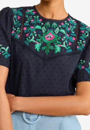 Blusa azul marino con mangas cortas abullonadas, con bordados florales verdes y rosas en el pecho y las mangas, combinada con pantalones azul claro.