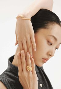 Pulsera en forma de serpiente de oro y anillo texturizado llevados en los dedos, destacando los intrincados detalles y el acabado brillante sobre un fondo neutro.