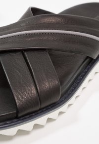 Sandal in pelle nera con cinturini incrociati, dotato di una suola zigzag bianca e cuciture grigie di accento lungo il cinturino. Finitura texturizzata.
