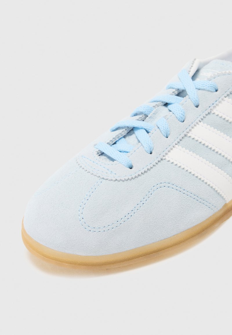 Hellblaue Wildledersneaker mit weißen Streifen und Schnürsenkeln, die ein strukturiertes Obermaterial und eine tanfarbene Gummisohle vor einem schlichten Hintergrund präsentieren.