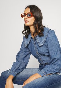 Camisa de denim com um grande colar franzido, tecido azul, mangas longas e bufantes, combinada com jeans azuis a condizer e óculos de sol retangulares laranja.