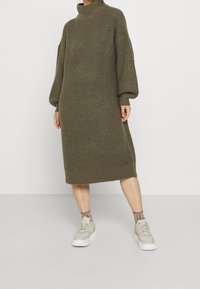 Olivgrön stickad klänning med hög krage, långa ärmar och ribbad textur, kombinerad med ljusa sneakers och ankelsockor.