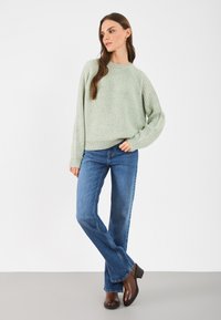 Gai zaļš adīts džemperis ar brīvu piegriezumu, apaļu kakla izgriezumu, garām piedurknēm; ap kombinēts ar ziliem platām džinsām un brūnām augstpapēžu potīšu zābakiem.