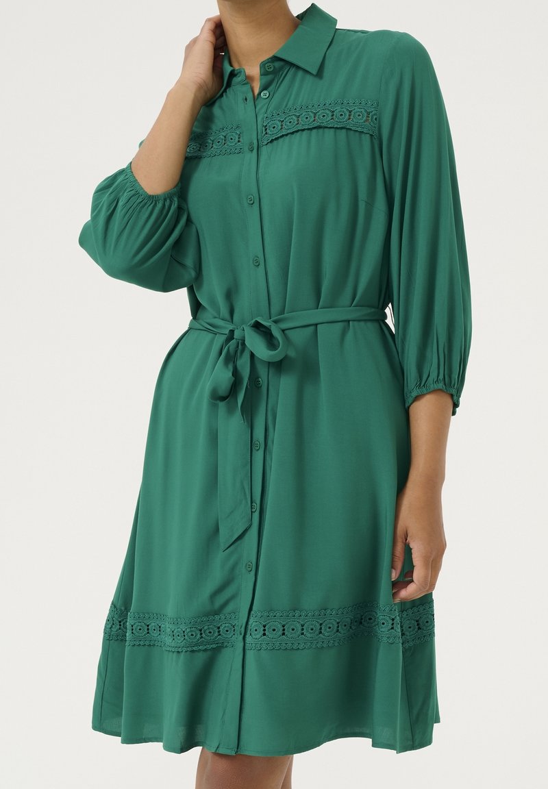 Femme portant une robe-chemise verte boutonnée avec une bordure en dentelle sur la poitrine et l'ourlet, manches bouffantes 3/4 et une ceinture nouée à la taille.