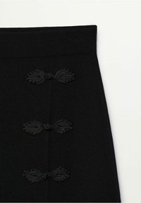 Jupe en maille noire avec une taille ajustée, ornée de trois accents décoratifs en boucle sur le devant. Texture lisse et design simple.