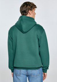 Zelen hoodies s prijetnim materijalom, velikim kapuljačom i rebrastim manžetama. Stražnja strana je glatka bez vidljivih logotipa ili uzoraka. Uparen s svijetloplavim trapericama.