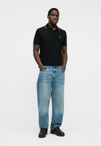 Άνδρας που στέκεται με τα χέρια στις τσέπες, φοράει μαύρο πόλο πουκάμισο, ανοιχτό μπλε τζιν και μαύρα παπούτσια μπροστά σε απλό λευκό φόντο.