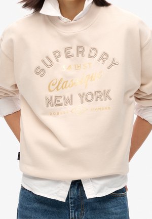 Osoba nosící béžovou mikinu s nápisem "Superdry New York 34th St Classique Double Diamond", bílou košili pod ní a modré džíny.