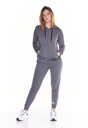 Une femme porte un sweat à capuche gris zippé et un pantalon de jogging assorti, avec des baskets blanches, sur un fond blanc uni.