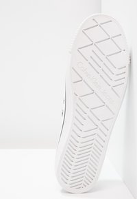 Weißer Sneaker mit strukturiertem Gummisohle, der ein Profilmuster, metallische Ösen und die Gravur "Calvin Klein Jeans" an der Seite aufweist.