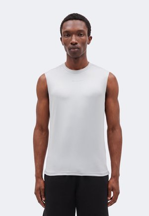 Homme aux cheveux courts portant un débardeur de sport gris clair et un short noir, debout devant un fond clair uni.