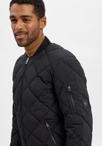 Veste bomber matelassée noire avec un col côtelé, des poches latérales zippées et un patch logo sur la manche. Texture lisse et design structuré.
