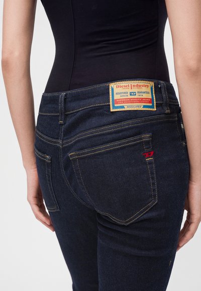 Personne portant un jean Diesel bleu foncé avec une étiquette à la taille et une couture rouge sur la poche arrière, vue de la taille vers le bas.