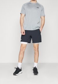 Camisa deportiva gris claro de manga corta con un logo negro, combinada con pantalones cortos negros que tienen una capa blanca por debajo y zapatillas negras.