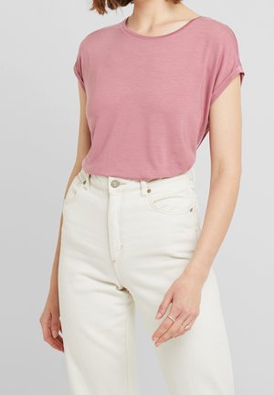 Kvinde iført en støvet lyserød t-shirt med korte ærmer, stoppet ned i højttaljede off-white bukser, med ringe på højre hånd.