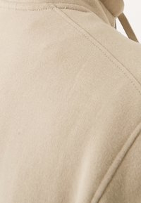 Gros plan sur la couture de l'épaule d'un sweat à capuche en tissu beige et détail du cordon de la capuche.