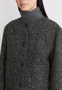 Grau gemusterte Tweedjacke mit Fischgrätmuster, ausgestattet mit einem hohen gerippten Kragen, fünf schwarzen Knöpfen und zwei vorderen Taschen.