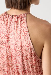 Blouse sans manches rose avec des motifs floraux rouges et abstraits, dotée d'un col froncé et d'une fermeture à bouton au dos.