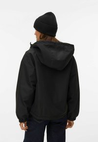 Kvinde set bagfra iført en sort hættetrøje, sort strikket hue og mørkeblå jeans mod en ensfarvet lys baggrund.