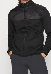 Mężczyzna w czarnej kurtce zapinanej na zamek z pikowanym panelem na górnej części klatki piersiowej i logo "CK", ręce w kieszeniach, zestawiony z ciemnymi spodniami.