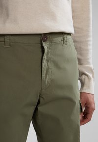 Persona vistiendo una blusa de manga larga beige claro metida dentro de unos pantalones verde oliva con cierre de botón y cremallera, con la mano relajada a un lado.