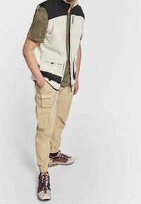Man in een kleurblokvest, olijfgroene shirt met korte mouwen, beige cargobroek met ketting en multicolore sneakers, staand tegen een witte achtergrond.